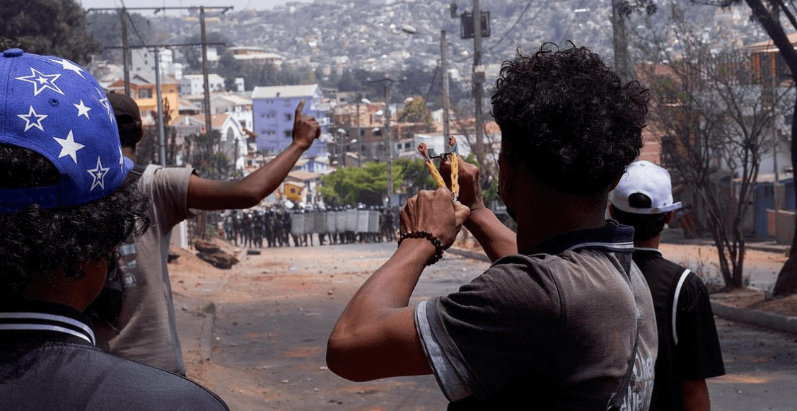 Varias personas jóvenes en una calle apuntan con hondas hacia una fila de policías antidisturbios al fondo.