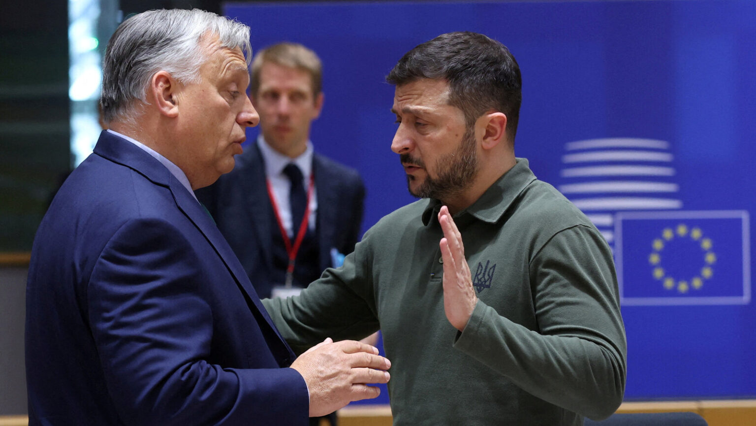 Viktor Orbán y Zelensky conversan durante una cumbre internacional, con la bandera de la Unión Europea al fondo.